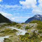 Scenic mountain landscape with a winding trail, lush greenery, and a blue sky dotted with clouds, showcasing the natural beauty of the outdoors.