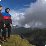 Two hikers standing on a grassy ridge, overlooking a mountainous landscape with clouds and blue sky, dressed in colorful outdoor jackets. Ideal for adventure travel and hiking content.