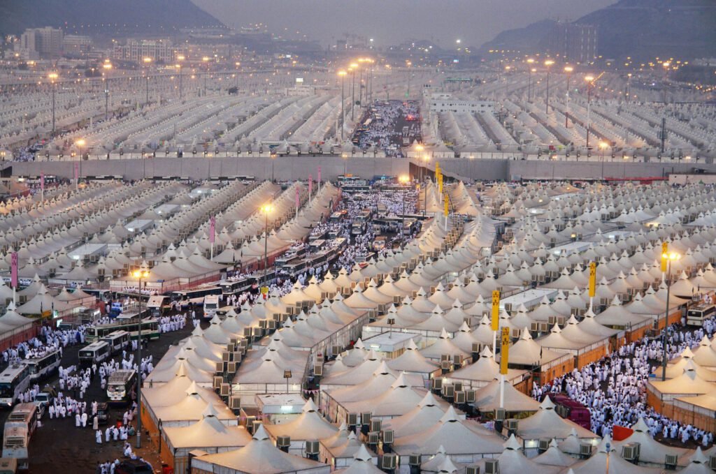 Aerial view of a large tent city at night, with numerous white tents arranged in rows, illuminated by lights, showcasing the scale of a significant event or gathering.