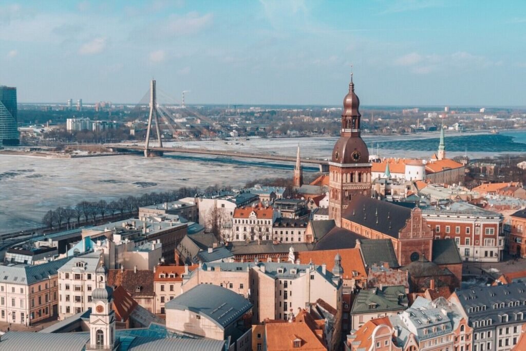 Aerial view of Riga, Latvia showcasing the historic architecture, including St. Peter's Church and the National Library, with the Daugava River in the background and a clear blue sky.