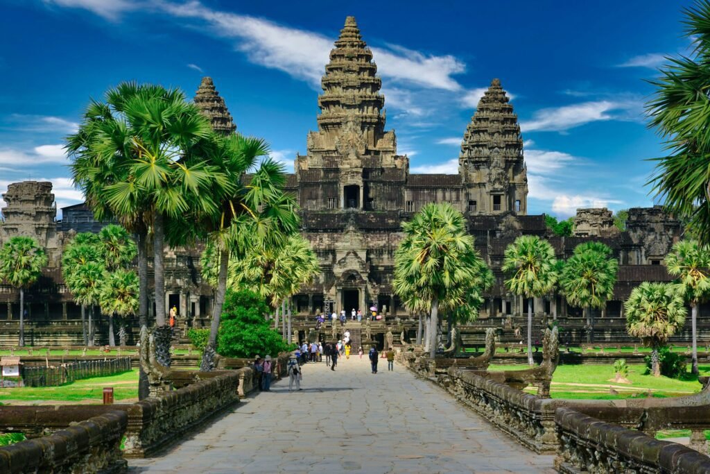 Angkor Wat temple complex in Cambodia, showcasing intricate architecture and lush palm trees under a clear blue sky. Visitors stroll along the pathway leading to the iconic towers.