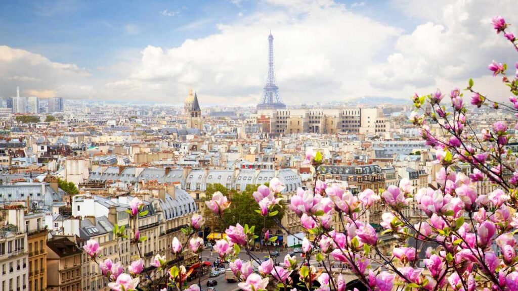 Panoramic view of Paris with the Eiffel Tower in the background, framed by blooming cherry blossoms under a partly cloudy sky.