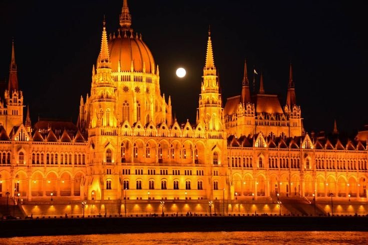 Illuminated Hungarian Parliament Building at night with a full moon in the background, showcasing its stunning Gothic architecture and vibrant orange lights reflecting on the water.