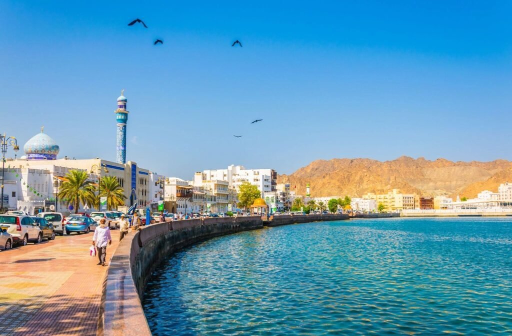 Scenic view of a coastal promenade in Muscat, Oman, featuring clear blue waters, modern buildings, and a mountainous backdrop under a bright blue sky. People stroll along the waterfront, with birds flying overhead.