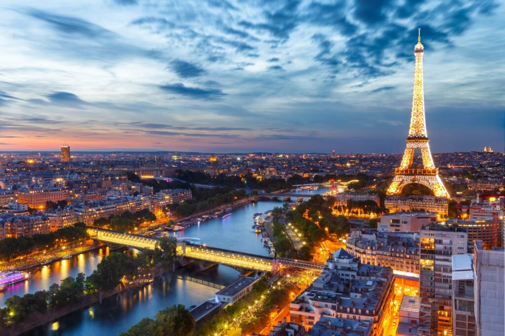 Panoramic view of Paris at dusk, featuring the illuminated Eiffel Tower and the Seine River, with vibrant city lights and a dramatic sky.