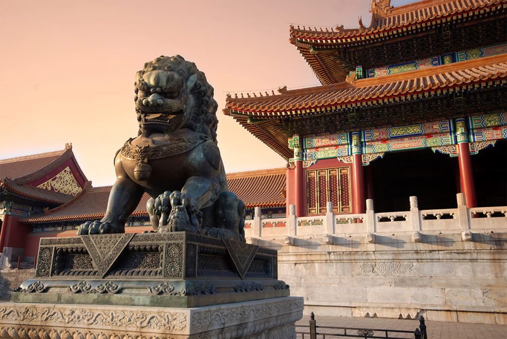 Bronze guardian lion statue in front of traditional Chinese architecture at sunset, showcasing intricate details and cultural significance.