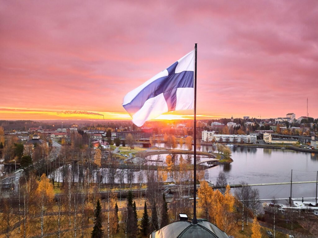 Finnish flag waving against a vibrant sunset over a scenic cityscape, showcasing the natural beauty and urban landscape of Finland.