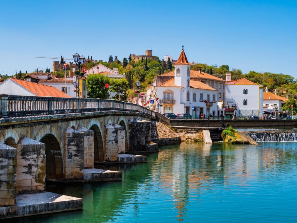Scenic view of a historic bridge over calm waters, with charming white buildings and a castle in the background under a clear blue sky.