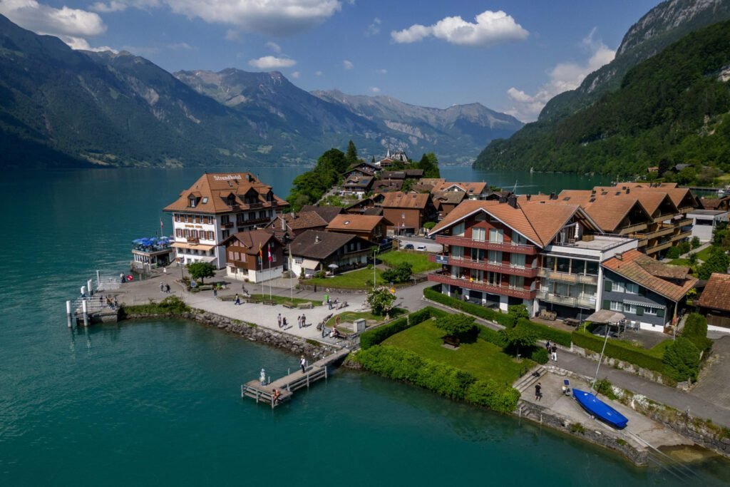 Aerial view of a picturesque Swiss village by a serene lake, surrounded by mountains, featuring traditional wooden chalets and lush greenery.