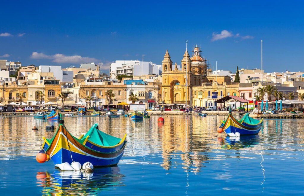 Colorful traditional Maltese fishing boats in the foreground, reflecting in the calm waters, with historic buildings and a clear blue sky in the background.