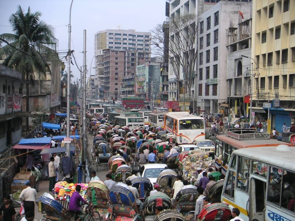 Bustling urban street scene in a densely populated city, showcasing heavy traffic congestion with various vehicles, including buses and rickshaws, amidst tall buildings and shops.