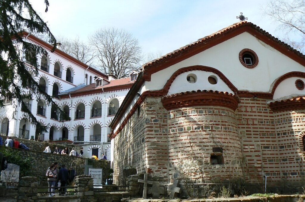 Historic monastery architecture featuring a stone building with a red-tiled roof and arched windows, surrounded by trees and visitors. The structure showcases traditional design elements typical of religious sites.