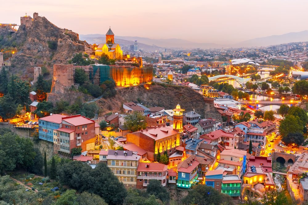 A panoramic view of Tbilisi, Georgia, showcasing the vibrant rooftops of the old town, the historic Narikala Fortress illuminated at dusk, and the scenic landscape of the surrounding hills.