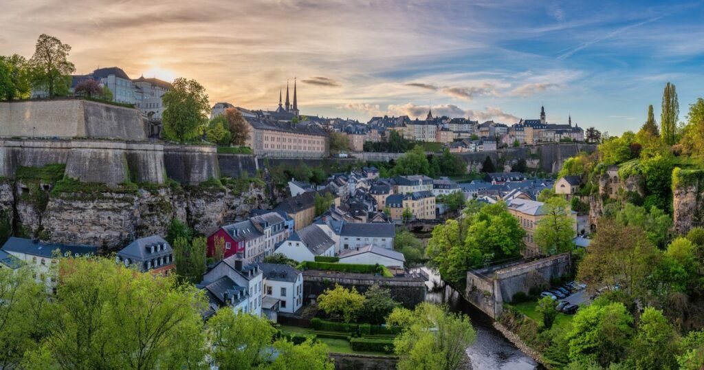 Panoramic view of Luxembourg City at sunset, showcasing historic architecture, lush greenery, and winding streets. The skyline features prominent church spires and elevated buildings, highlighting the city's blend of natural beauty and urban charm.
