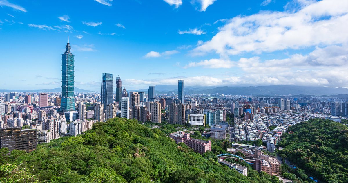 Panoramic view of a vibrant city skyline surrounded by lush greenery under a clear blue sky, showcasing modern skyscrapers and urban development.