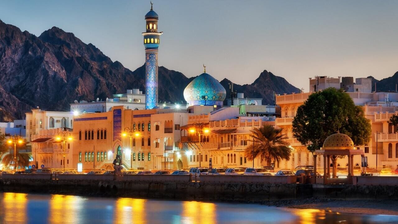 Scenic view of a coastal city at dusk, featuring a prominent mosque with a blue dome and minaret, surrounded by mountains and illuminated buildings along the waterfront.