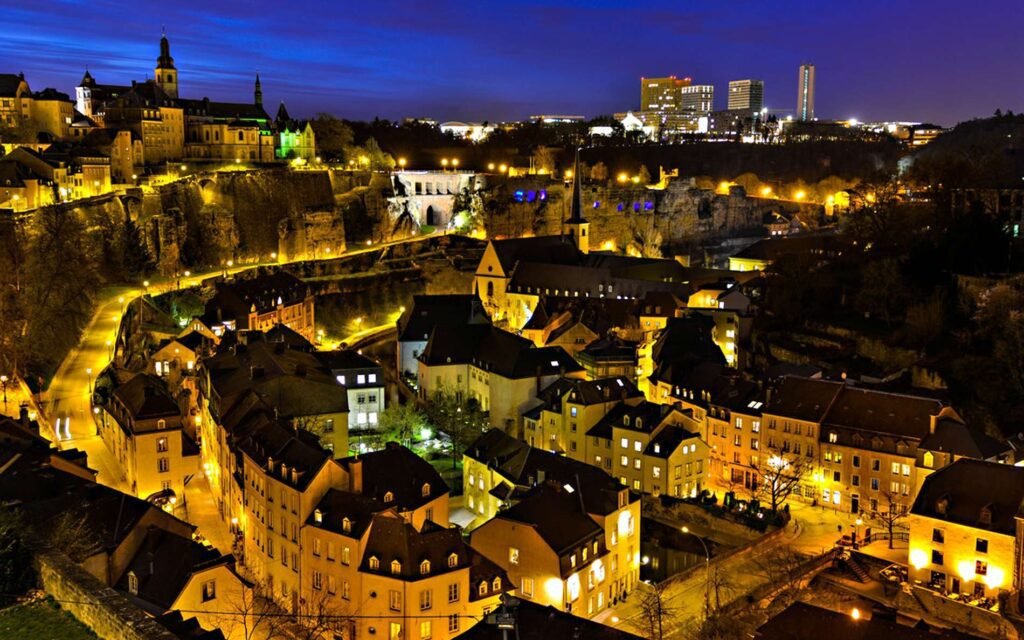 Aerial view of Luxembourg City at night, showcasing illuminated historic buildings and modern skyscrapers against a twilight sky.