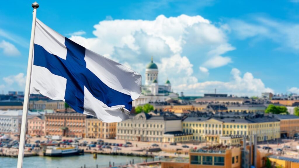Finnish flag waving in front of a scenic view of Helsinki, showcasing historic architecture and a cloudy sky.
