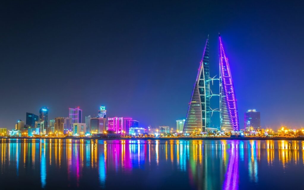 City skyline at night featuring illuminated skyscrapers, including the iconic Bahrain World Trade Center, with vibrant colors reflecting on the water.