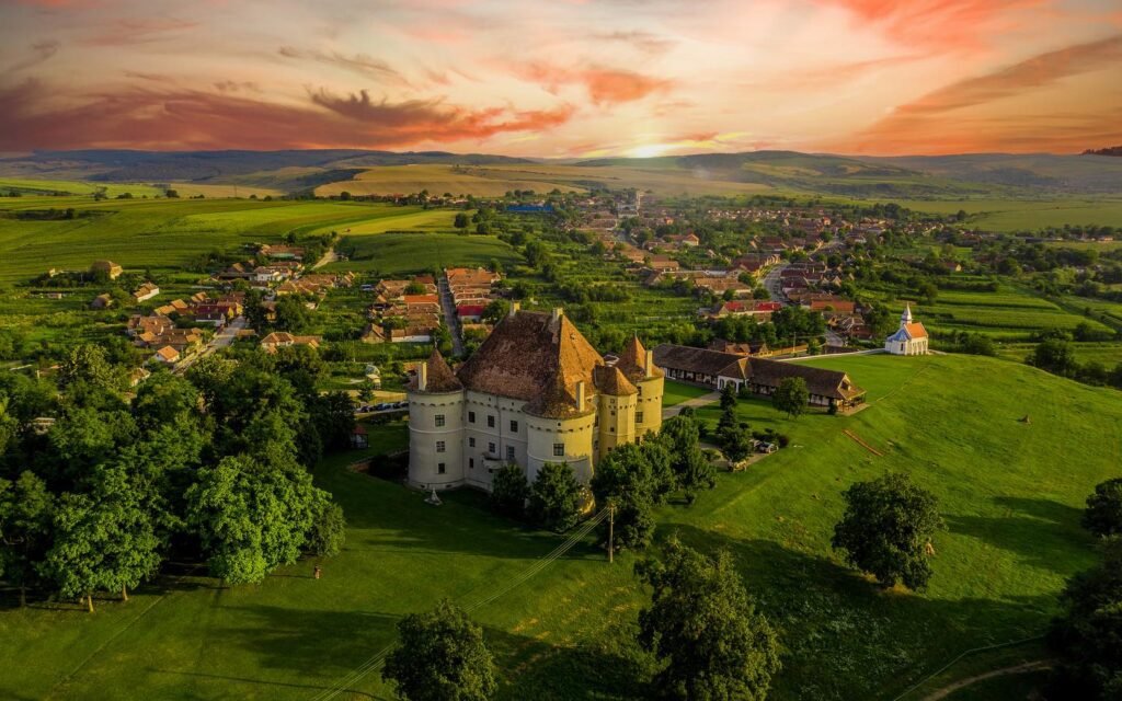 Aerial view of a picturesque landscape featuring a historic castle surrounded by lush green fields and quaint village homes under a vibrant sunset sky.