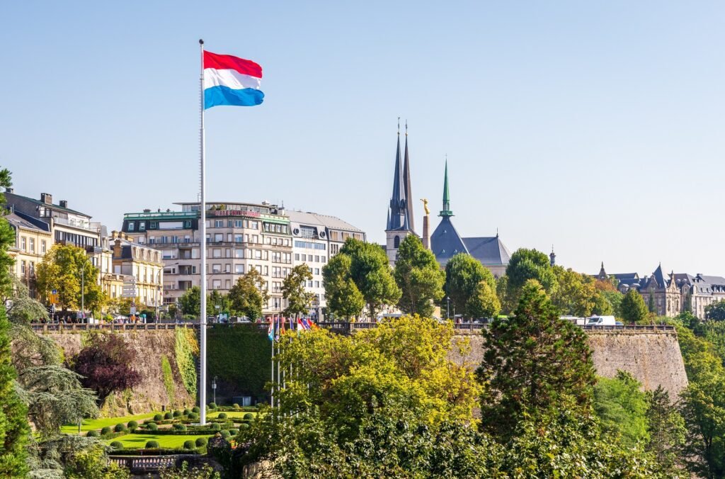 Luxembourg flag waving in the foreground with a scenic view of the city skyline, featuring historic spires and lush greenery under a clear blue sky.