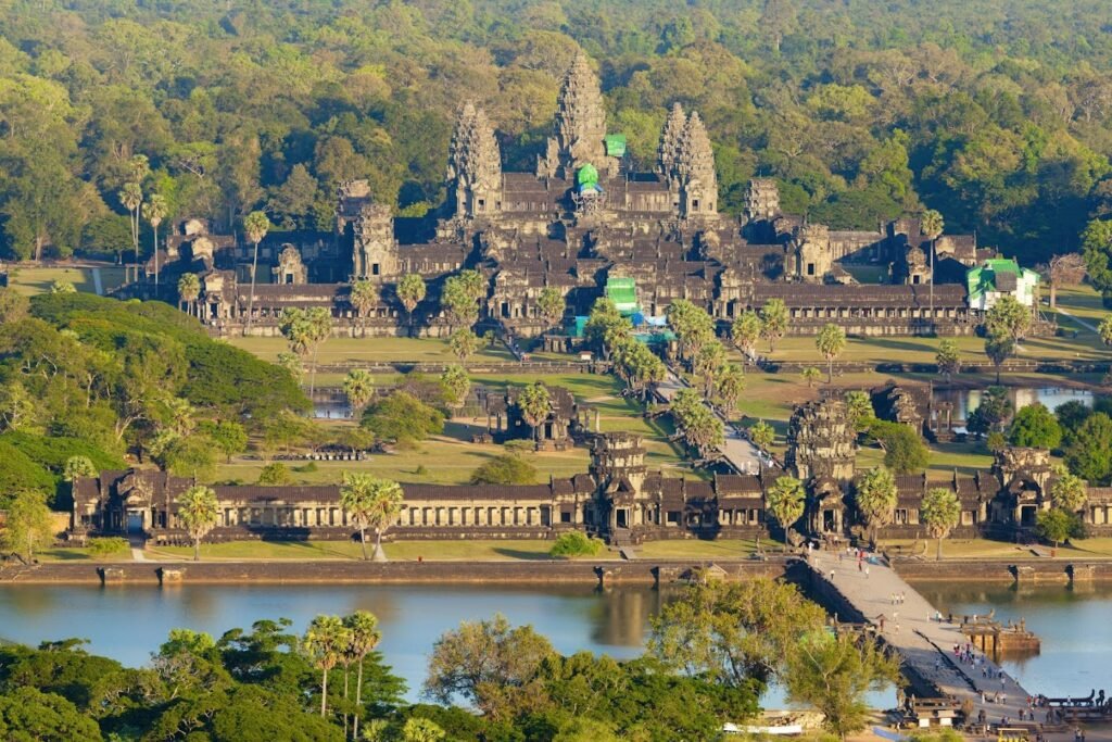 Aerial view of Angkor Wat, a UNESCO World Heritage Site in Cambodia, surrounded by lush greenery and a reflective water body, showcasing its intricate architecture and historical significance.