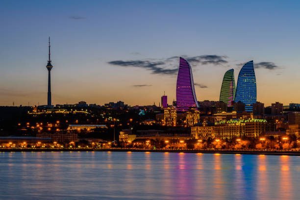 Baku skyline at dusk featuring the Flame Towers illuminated in vibrant colors, with the Caspian Sea in the foreground and the TV tower visible on the left.