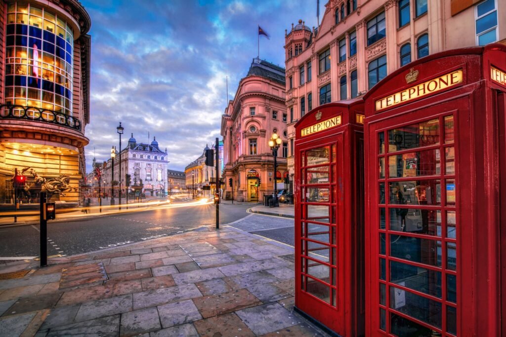Red telephone boxes on a cobblestone street in London at dusk, with historic buildings and a dramatic sky in the background.