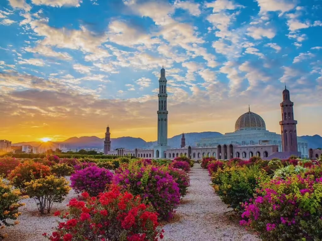 Scenic view of a mosque at sunset surrounded by vibrant flowers and mountains, with a colorful sky and a prominent minaret in the background.