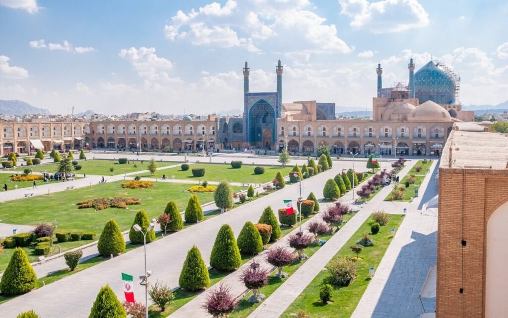 Aerial view of Naqsh-e Jahan Square in Isfahan, Iran, showcasing the lush gardens, symmetrical pathways, and historic architecture, including the Shah Mosque and Ali Qapu Palace under a bright blue sky.