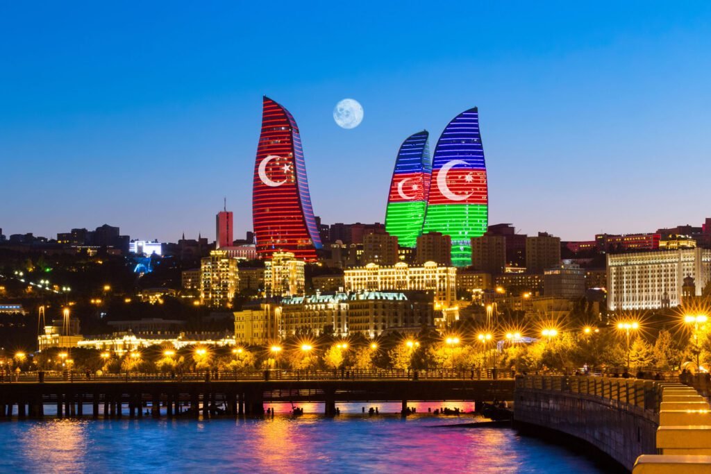 Baku's Flame Towers illuminated with the Turkish flag colors at dusk, showcasing a vibrant skyline against a moonlit sky. The image captures the architectural beauty and cultural significance of Azerbaijan's capital.