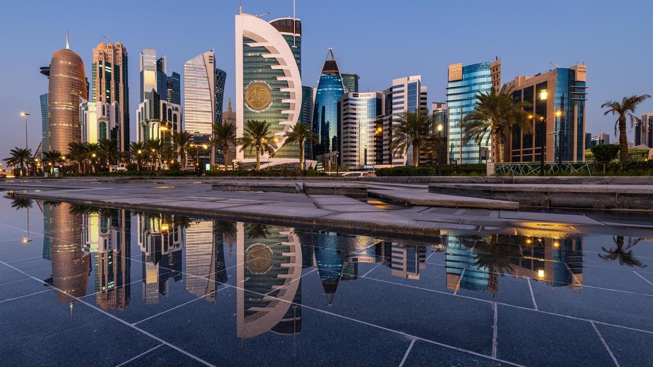 Cityscape of Doha, Qatar at dusk featuring modern skyscrapers and palm trees, with reflections in a water feature in the foreground.