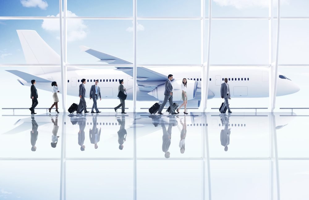 Business travelers walking through an airport terminal with an airplane visible in the background, reflecting a busy travel environment.
