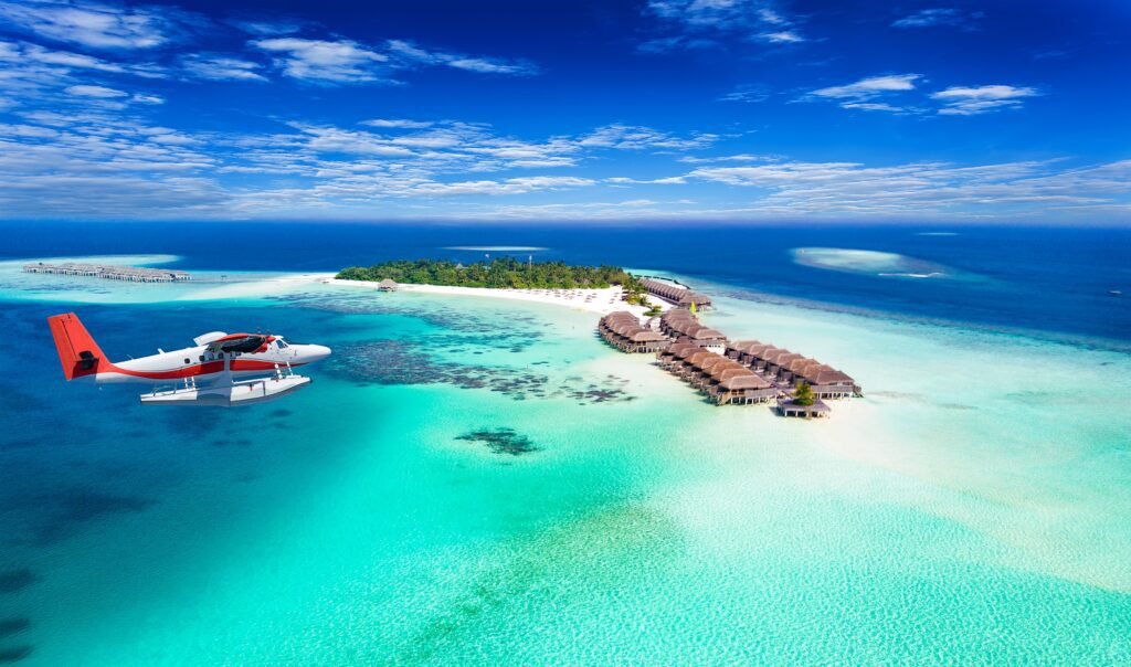Aerial view of a tropical island resort featuring overwater bungalows, clear turquoise waters, and a white sandy beach under a bright blue sky.