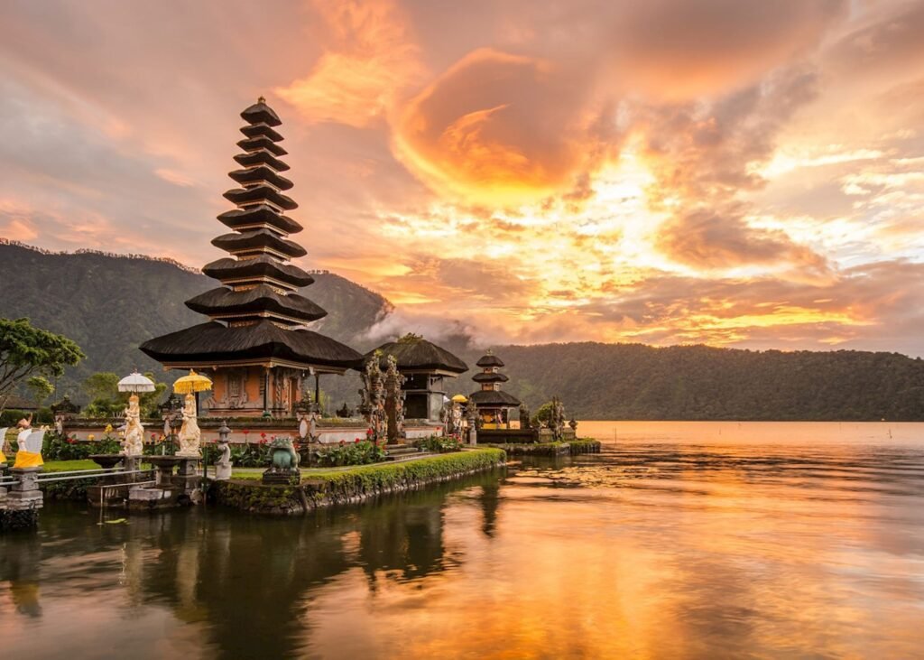 Sunset over Pura Ulun Danu Bratan temple in Bali, Indonesia, with a serene lake reflecting vibrant orange and purple skies. The iconic multi-tiered pagoda is surrounded by lush greenery and mountains in the background.