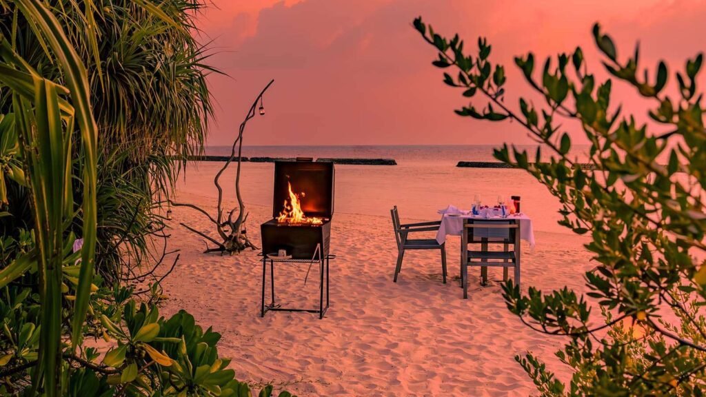 Beach barbecue setup at sunset with a grill and dining table on the sand, surrounded by tropical plants and a serene ocean view.