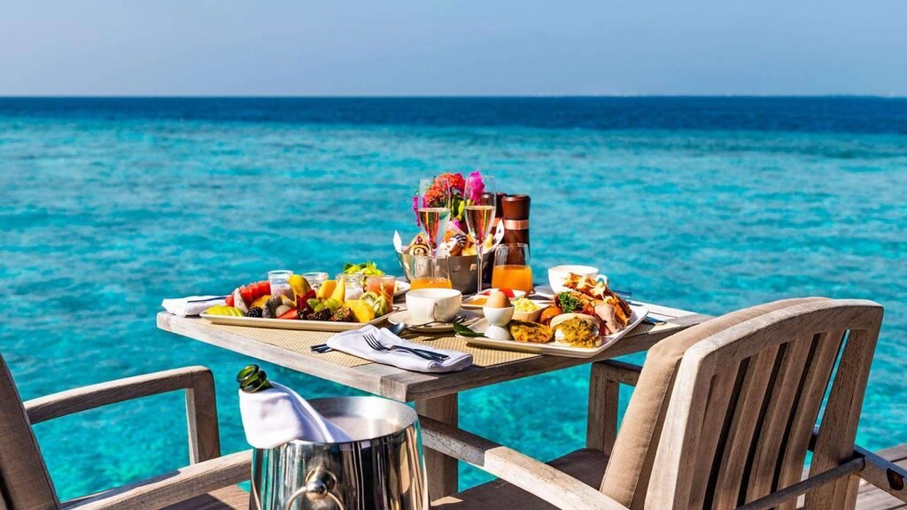 Luxurious beachside dining setup featuring a variety of gourmet dishes, fresh fruits, and beverages on a wooden table with a stunning ocean view in the background. Perfect for a tropical getaway or romantic meal.