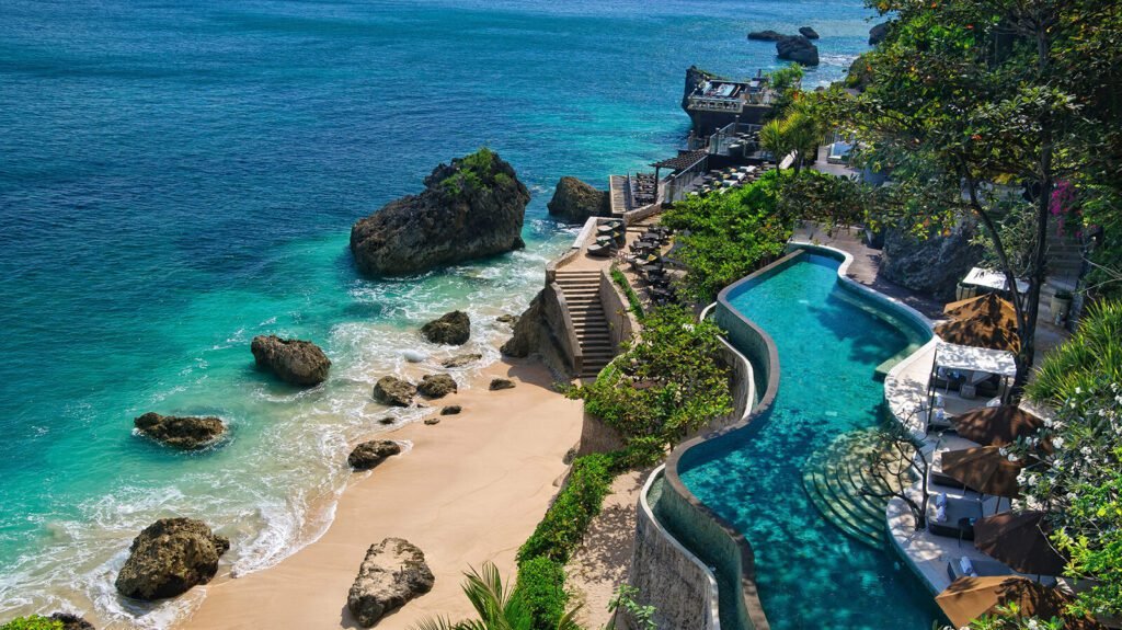 Scenic view of a luxurious beachfront resort featuring a winding infinity pool, sandy beach, and rocky shoreline, with crystal-clear turquoise waters and lush greenery surrounding the area.