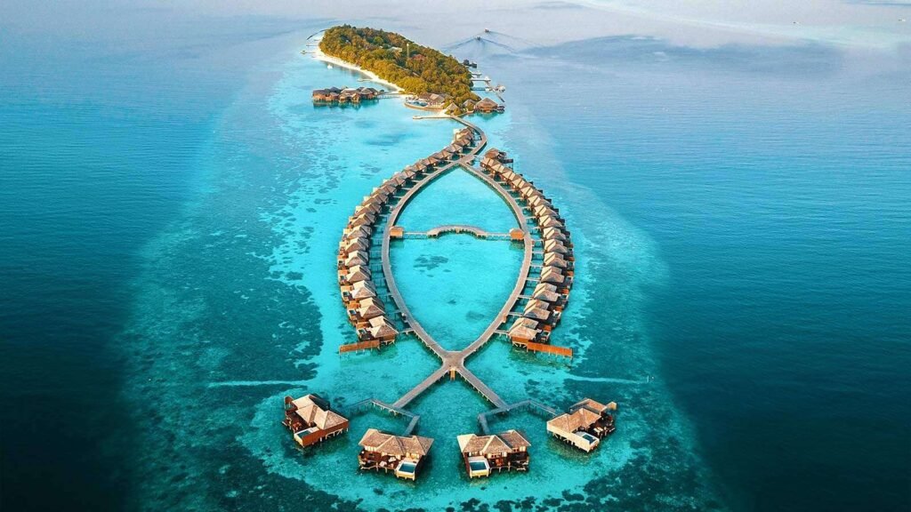 Aerial view of a luxury resort in the Maldives featuring overwater bungalows arranged in a unique fish shape, surrounded by vibrant turquoise waters and lush green island landscapes.