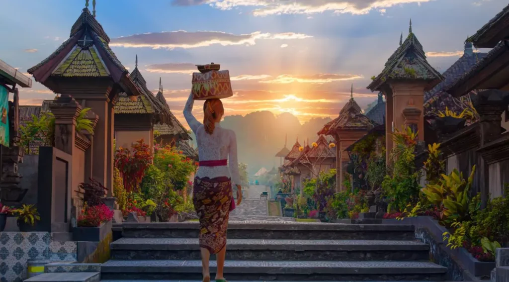Balinese woman carrying a traditional offering on her head, walking through a scenic temple pathway at sunset, surrounded by lush greenery and ornate architecture.