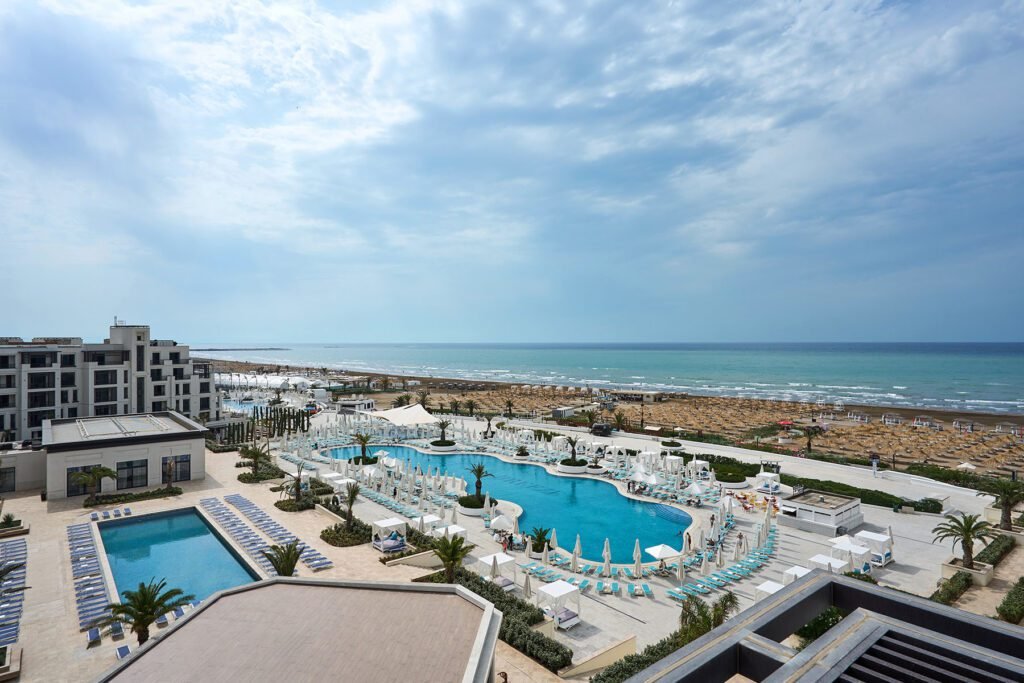 Aerial view of a luxurious beachfront resort featuring a large swimming pool, sun loungers, and palm trees, with the ocean and sandy beach visible in the background under a partly cloudy sky.