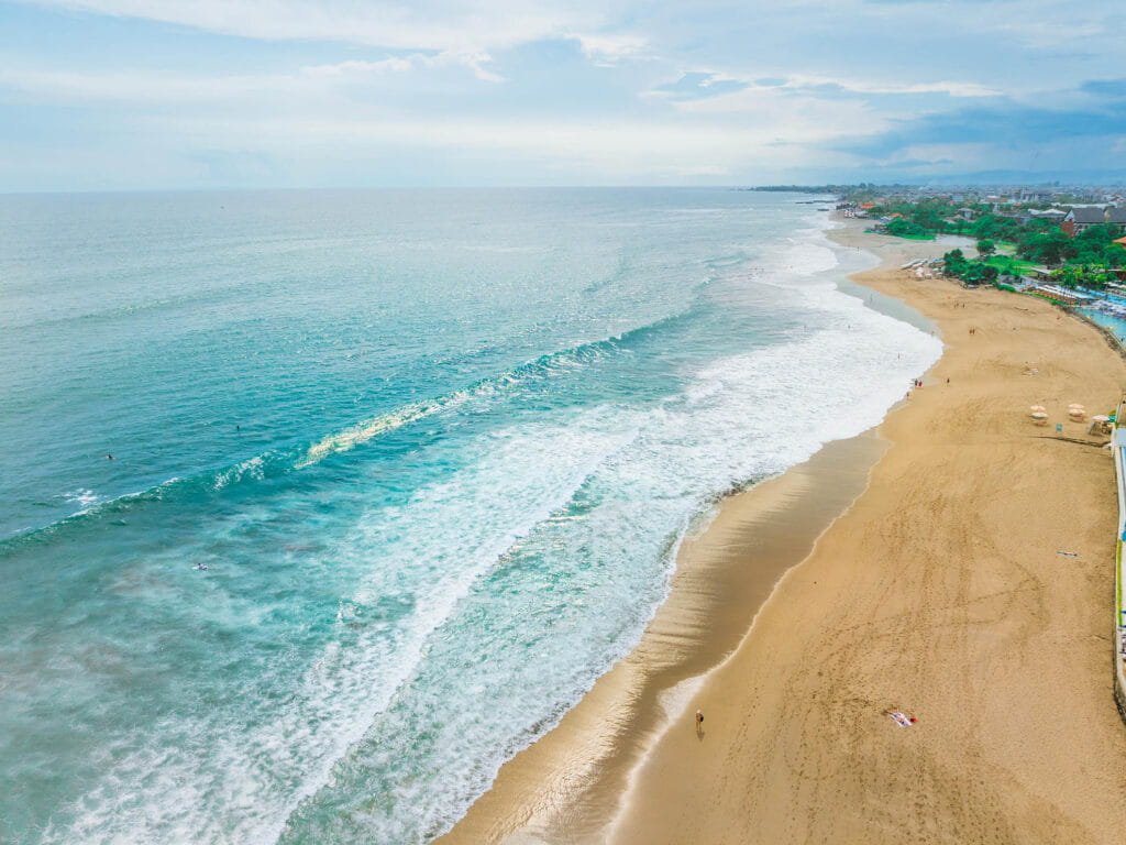 Aerial view of a serene beach with golden sand and gentle waves, showcasing a tranquil ocean scene under a partly cloudy sky, ideal for relaxation and outdoor activities.