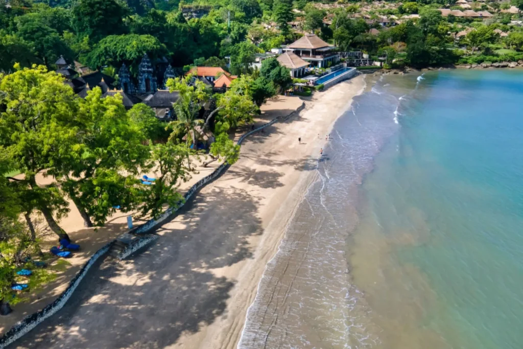 Aerial view of a serene beach with soft sand, lush green trees, and a clear blue ocean, featuring beachgoers walking along the shoreline and a resort with traditional architecture in the background.