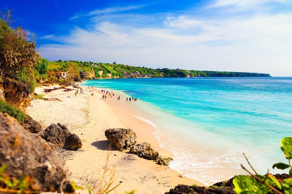Scenic view of a tropical beach with golden sand, turquoise waters, and lush green cliffs in the background, featuring people enjoying the sun and swimming. Ideal destination for relaxation and outdoor activities.
