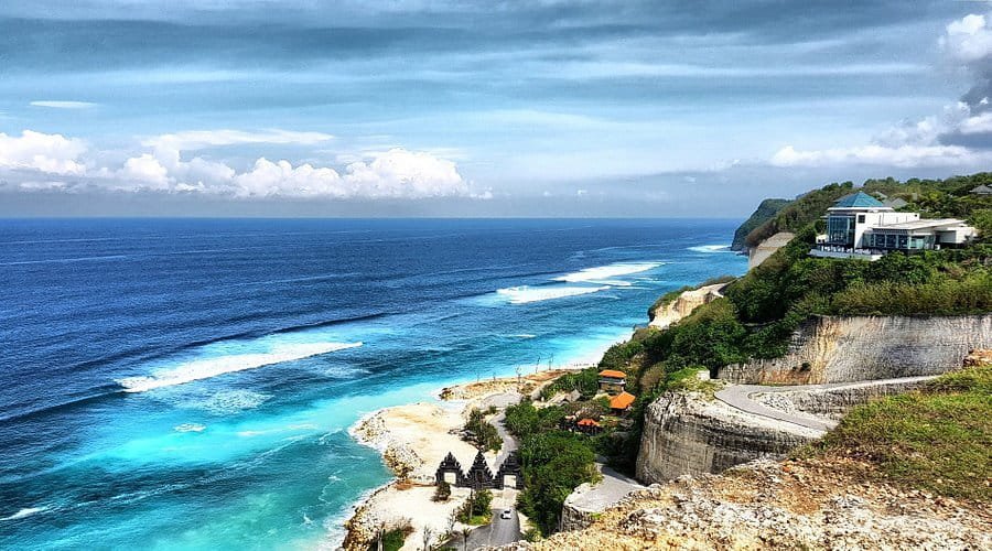 Scenic coastal view of turquoise waters meeting the blue sky, featuring cliffs and a winding road along the shoreline, with lush greenery and modern architecture in the background. Ideal for showcasing Bali's natural beauty and travel destinations.