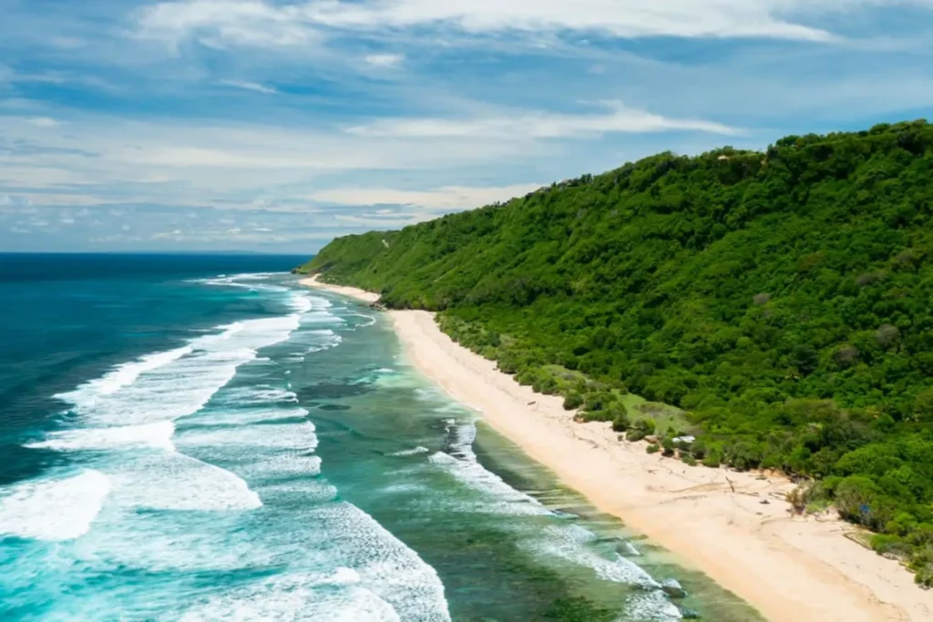 Aerial view of a pristine beach with turquoise waters and gentle waves, bordered by lush green hills under a bright blue sky. Ideal for tropical vacation and nature photography.