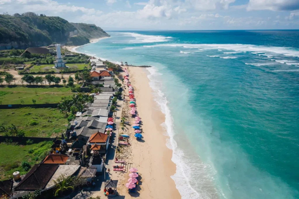 Aerial view of a beautiful beach in Bali, featuring colorful umbrellas lining the sandy shore, lush greenery, and a lighthouse in the background. The clear turquoise waters gently lap against the shoreline, creating a picturesque tropical scene perfect for vacationers.