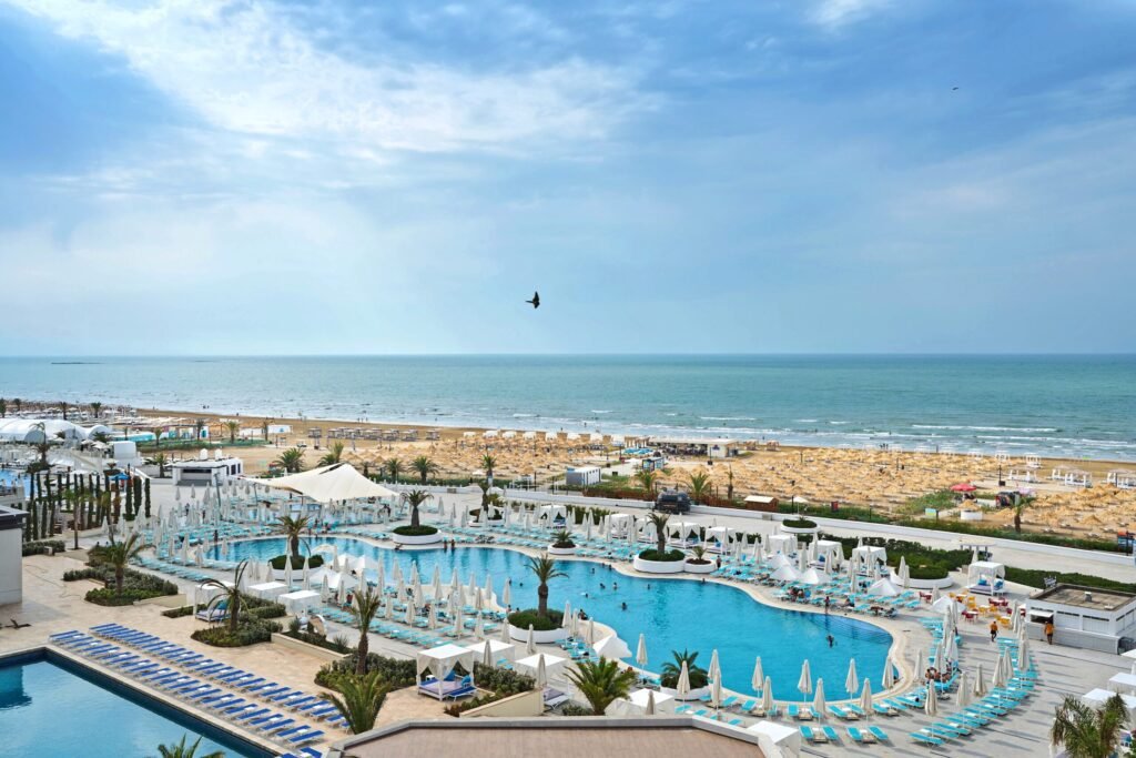 Panoramic view of a luxurious beach resort featuring a large swimming pool surrounded by sun loungers, palm trees, and cabanas, with a sandy beach and the ocean visible in the background under a partly cloudy sky.