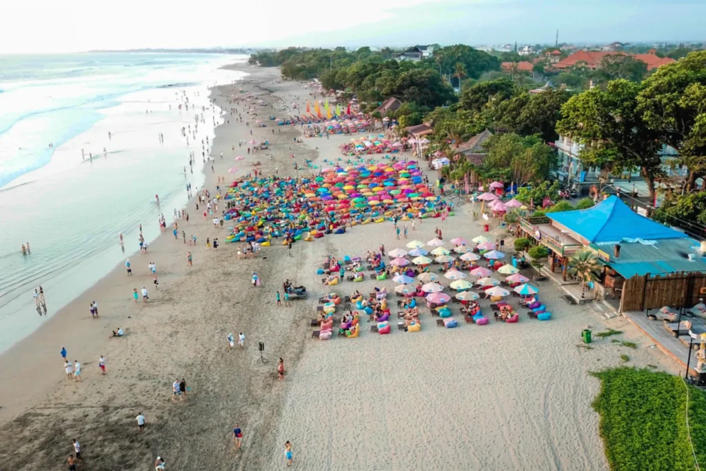 Aerial view of a bustling beach with colorful umbrellas and sun loungers, filled with people enjoying the sun and waves, showcasing a vibrant seaside atmosphere.