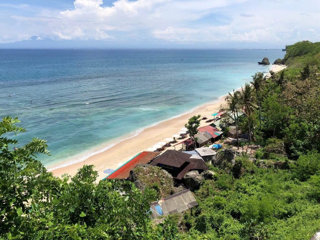 Scenic view of a tropical beach with clear blue water, sandy shore, and lush greenery. Beachside huts and sun loungers are visible under palm trees, with distant mountains on the horizon. Ideal destination for relaxation and water activities.